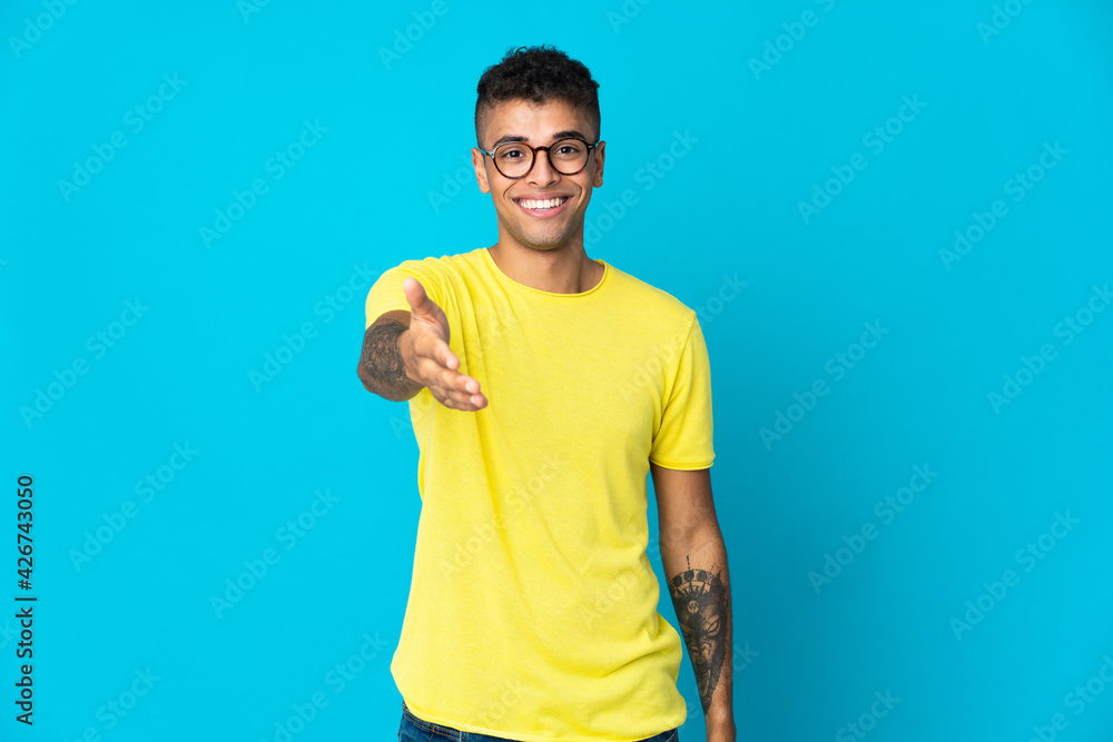 Young Brazilian man isolated on blue background shaking hands for closing a good deal Young Brazilian man isolated on blue background shaking hands for closing a good deal