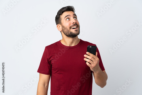 Young handsome man with beard using mobile phone isolated on white background looking up while smiling