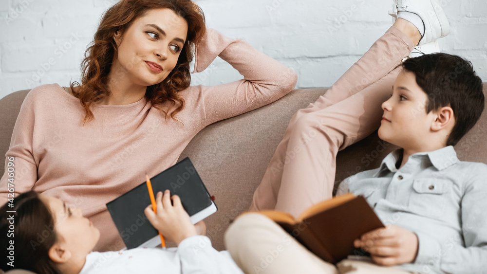 Woman looking at kids with notebook and book on blurred foreground on couch. Woman looking at kids with notebook and book on blurred foreground on couch.