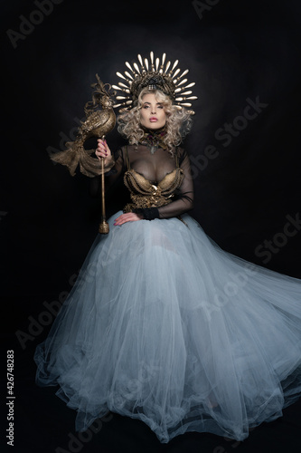 A gorgeous woman in a golden crown, the shape of the sun, a mythical creature, a queen. Photo in the studio. The concept of a fairy tale, legend, theater, cinema