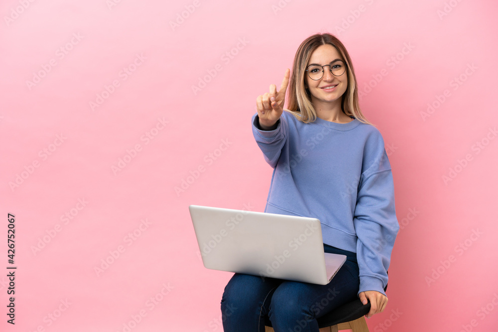 Fototapeta premium Young woman sitting on a chair with laptop over isolated pink background showing and lifting a finger