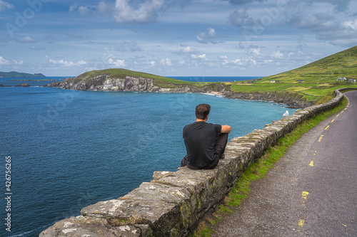 Middle age man sitting on a wall, admiring amazing view of Slea Head and Dingle Peninsula