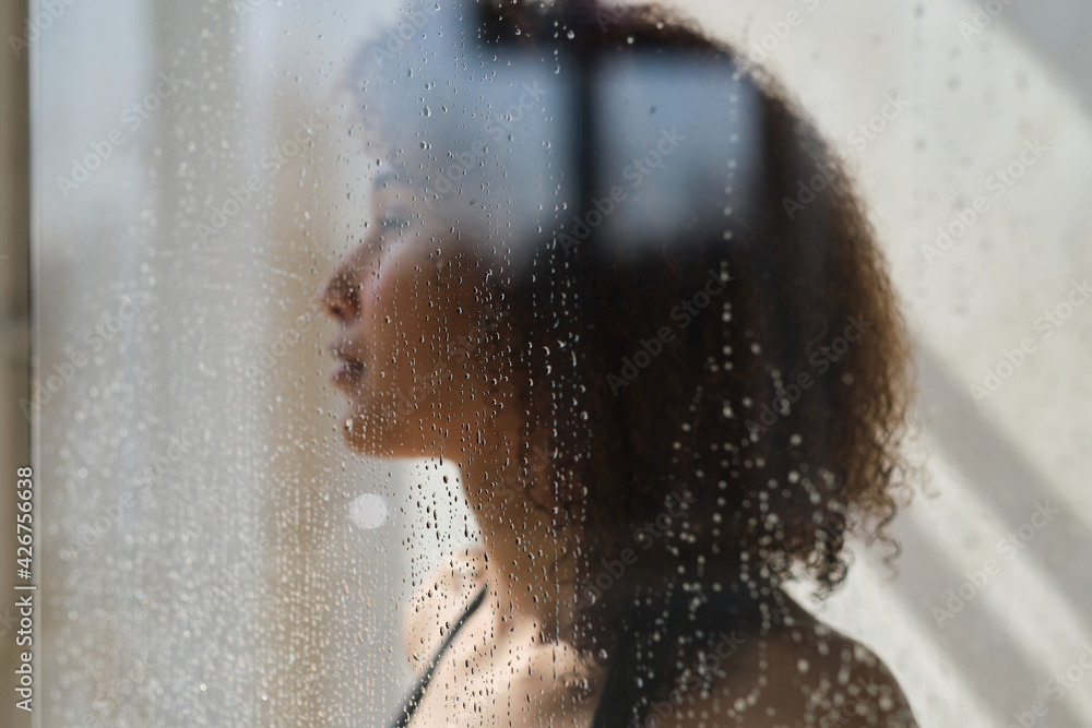 Sensual portrait of young woman taking a shower. Defocused female looks through the glass of the shower stall.