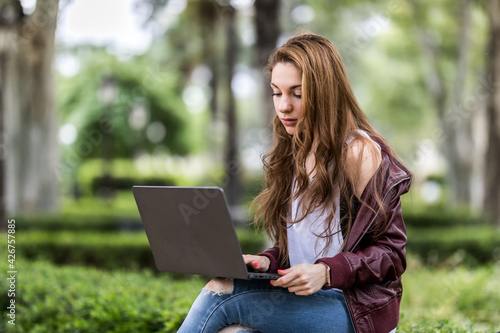 Young pretty woman works on laptop sitting on park bench near university close up