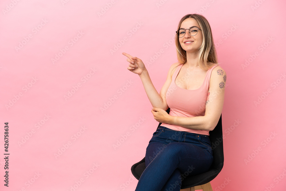 Young woman sitting on a chair over isolated pink background pointing finger to the side Young woman sitting on a chair over isolated pink background pointing finger to the side