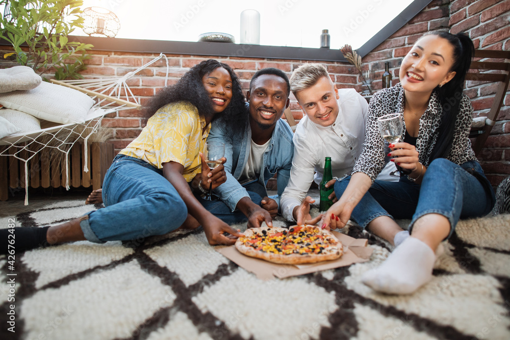 Four multicultural people in stylish clothes taking pieces of tasty pizza from box, smiling and looking at camera. Cheerful friends drinking alcoholic cocktails and eating snacks on party.
