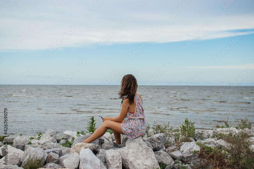 A beautiful young woman sits on a stone shore and looks at the ocean. Summer background. A beautiful young woman sits on a stone shore and looks at the ocean. Summer background.