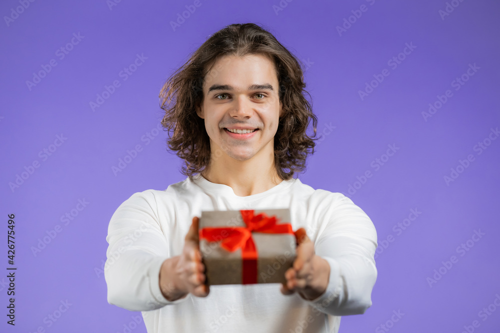 Charming man gives gift box by hand to camera on violet wall background. Guy smiling, he is happy with present. Studio portrait