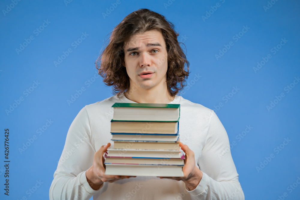 European student is dissatisfied with amount of homework and books on blue background. Man with long hairdo in displeasure, he is annoyed,discouraged frustrated by studies European student is dissatisfied with amount of homework and books on blue background. Man with long hairdo in displeasure, he is annoyed,discouraged frustrated by studies