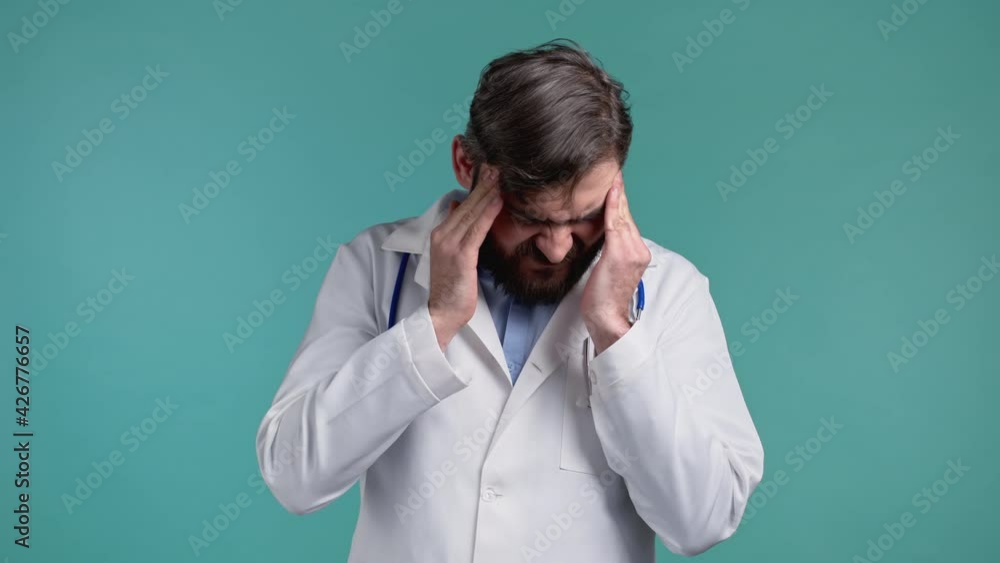Doctor man having headache, studio portrait. Bearded doc putting hands on head, isolated on blue background. Concept of problems, medicine, illness Doctor man having headache, studio portrait. Bearded doc putting hands on head, isolated on blue background. Concept of problems, medicine, illness
