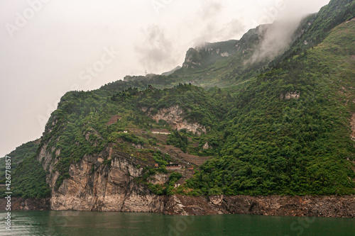 Yangtze River, Xiling Gorge, China - May 6, 2010: Xiangxicun region. Landscape, Agriculture on terraces on slope of green forested mountains under silver foggy sky. Farm houses and green water.