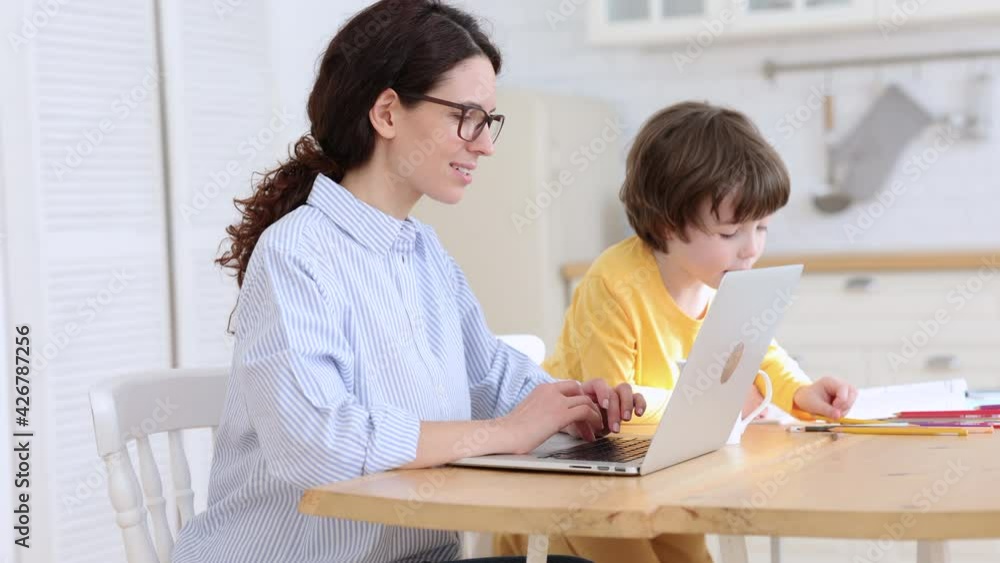 Family, lockdown distance job concept. Kid son painting while his mother is working on laptop at home office. Woman with her child doing homework or studying sitting together at table in living room. Family, lockdown distance job concept. Kid son painting while his mother is working on laptop at home office. Woman with her child doing homework or studying sitting together at table in living room.