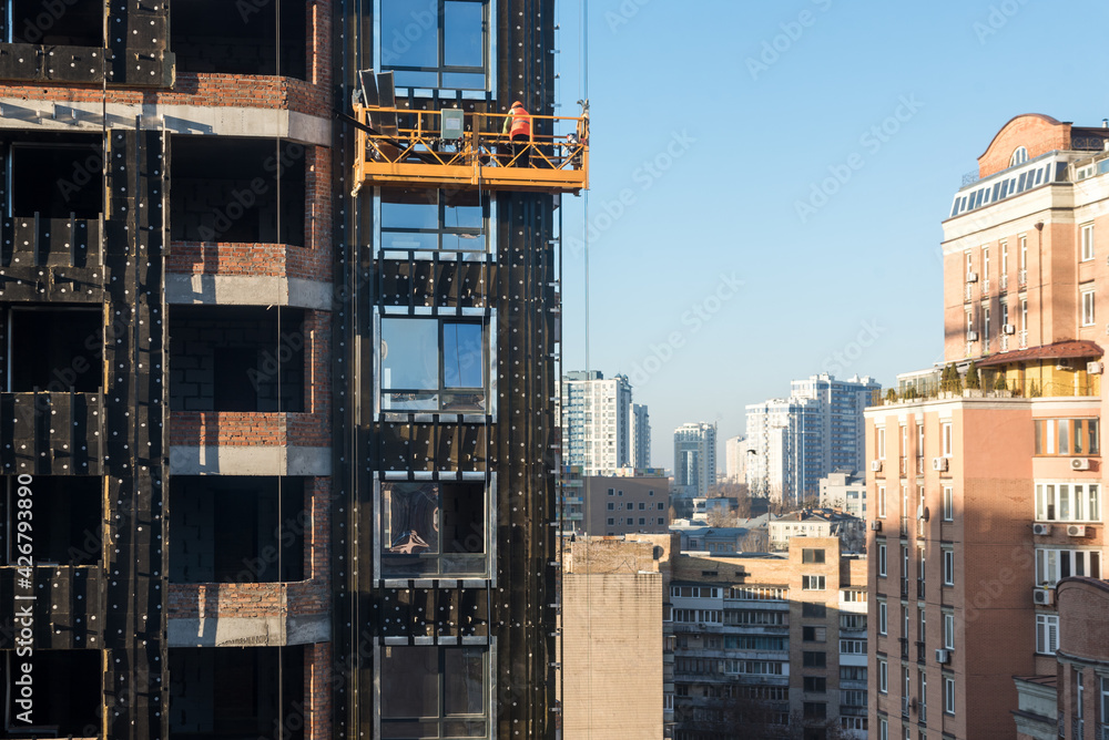 Fototapeta premium High rise construction work. Construction site workers in cradles working with facade. Suspended cradle for builders to work outside the skyscraper. Building construction