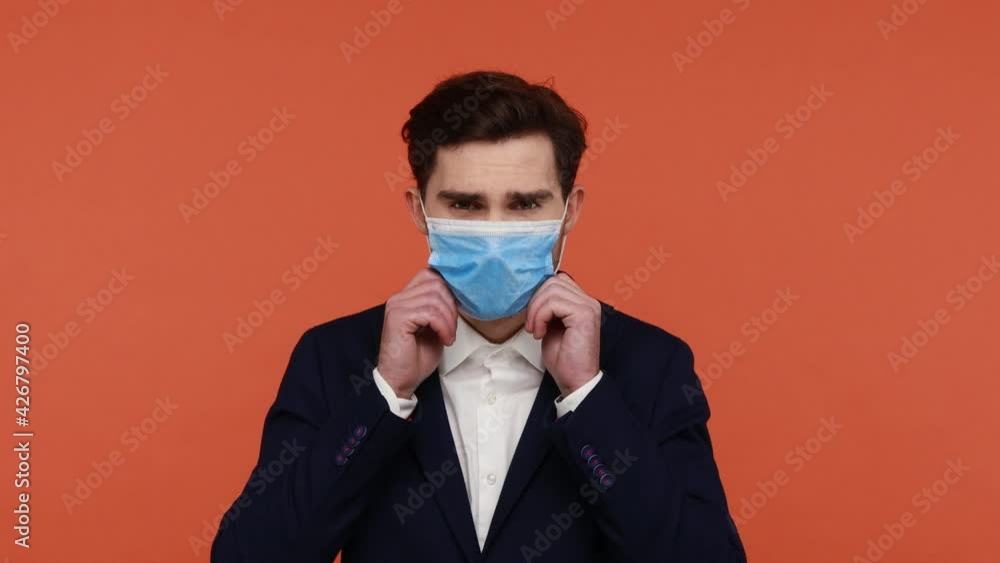 Young bearded handsome guy puts on and then takes off the medical mask, protection from virus, prevents the spread of covid, invoke to care of safety. Indoor studio shot isolated on orange background Young bearded handsome guy puts on and then takes off the medical mask, protection from virus, prevents the spread of covid, invoke to care of safety. Indoor studio shot isolated on orange background