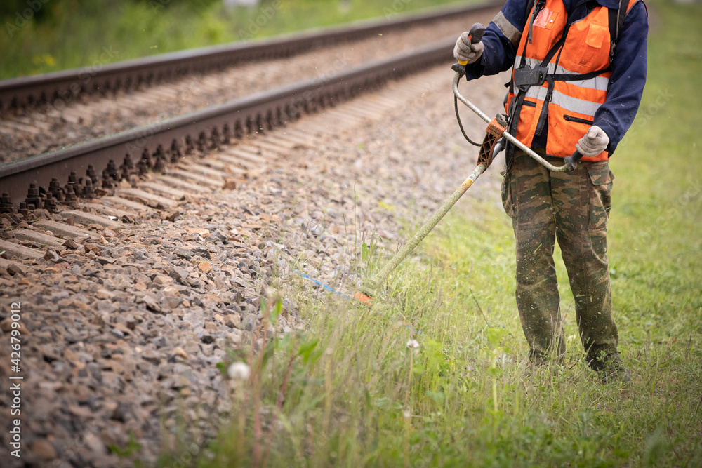 Obraz premium A worker in protective clothing walks along the lawn next to the train tracks and mows the grass with a gasoline lawn mower. A man mows the grass by the side of the road on a cloudy autumn day