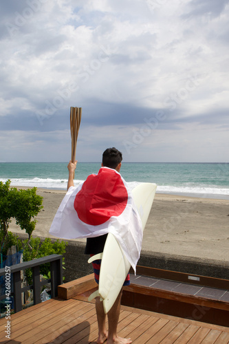 Man standing holding a flaming torch used in a large sporting event, with a surfboard and a Japanese Flag looking at the Pacific Ocean in Chiba Japan where a big surfing competition is held. photo