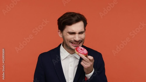 Bearded man wearing official style suit holding and smelling donut, harmful carbs, struggling temptation to eat sweet sugary doughnut. Indoor studio shot isolated on orange background