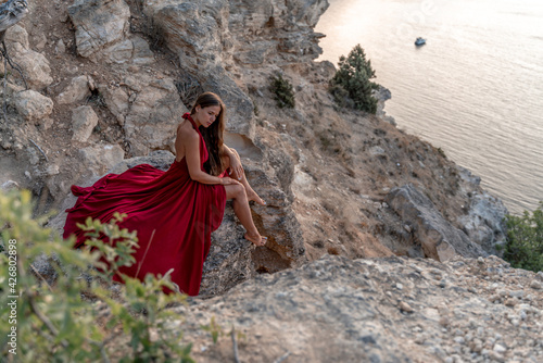 A girl with loose hair in a red dress sits on a rock rock above the sea. In the background  the sea. The concept of travel.