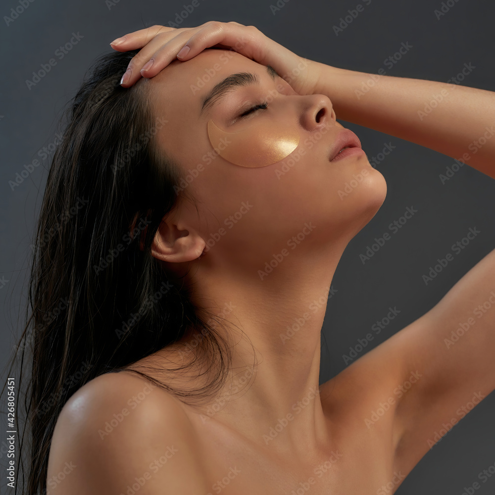 Naklejka premium Young beautiful asian half naked woman in bath towel with cosmetic eye patches touching her face while eyes closed