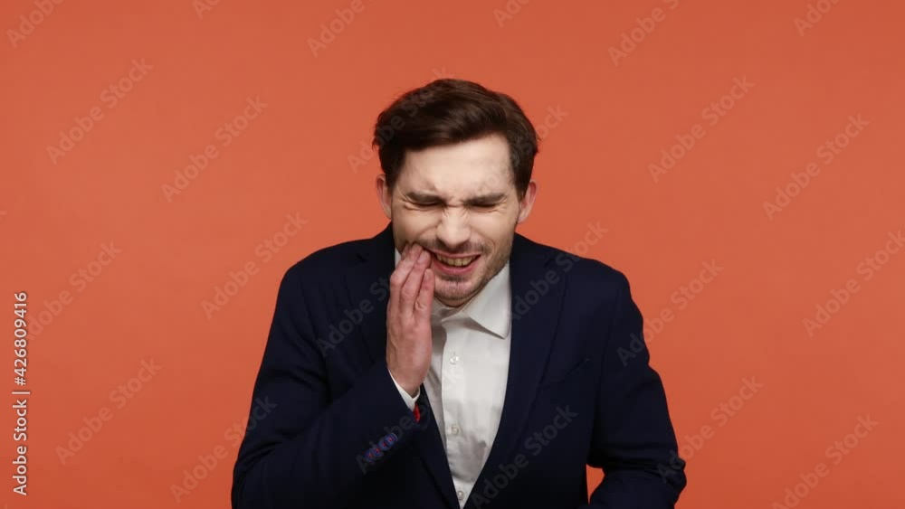 Young bearded man in official suit drinking hot or cold beverage and feeling unpleasant sore pain in teeth and high sensitivity, dental problems. Indoor studio shot isolated on orange background Young bearded man in official suit drinking hot or cold beverage and feeling unpleasant sore pain in teeth and high sensitivity, dental problems. Indoor studio shot isolated on orange background