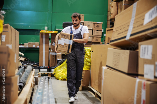Hardworking tattooed bearded manual worker walking trough warehouse and carrying very heavy big box ready for export.