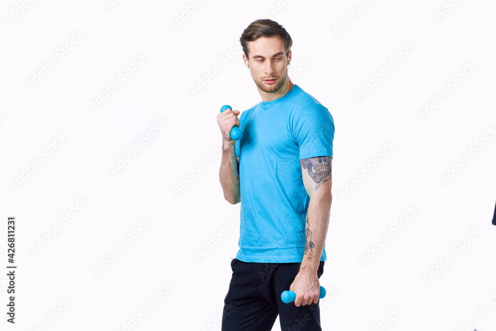 young man in a blue t-shirt with dumbbells on a light background and black pants fitness model young man in a blue t-shirt with dumbbells on a light background and black pants fitness model