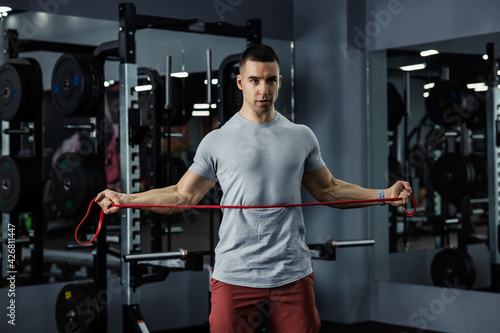 Professional male trainer doing fitness exercises with an elastic resistance band in a dark gym atmosphere. Handsome young man training with a rubber band. Sports life