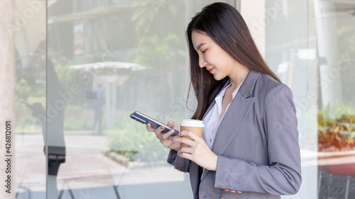 Young Asian woman wearing a gray suit is holding a phone and a paper coffee mug, Business women use smartphones to communicate by chatting or talking with business partners, Online media, VDO call.