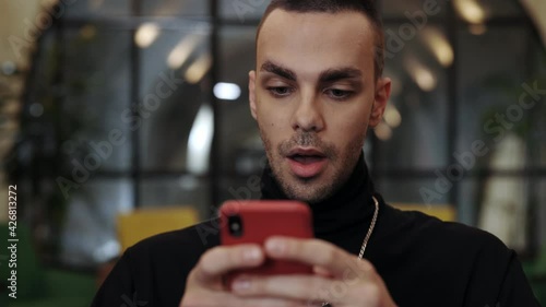 Close up view of caucasian man with bristles looking amazed and saying wow while using smartphone in cafe. Good looking guy with chainlet scrolling social media news feed and showing emotions. photo