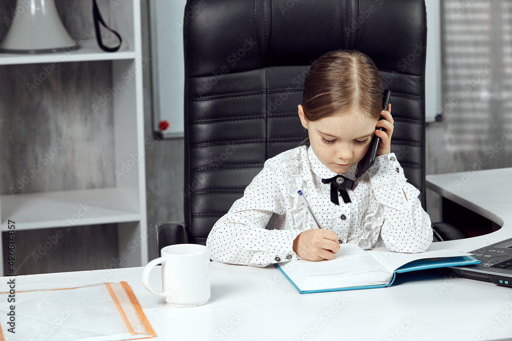 Portrait of a small cute child depicting the head of the agency at a white table, talking on a mobile phone. In the background is a bookcase with accounting records. The concept of adult children. Portrait of a small cute child depicting the head of the agency at a white table, talking on a mobile phone. In the background is a bookcase with accounting records. The concept of adult children.