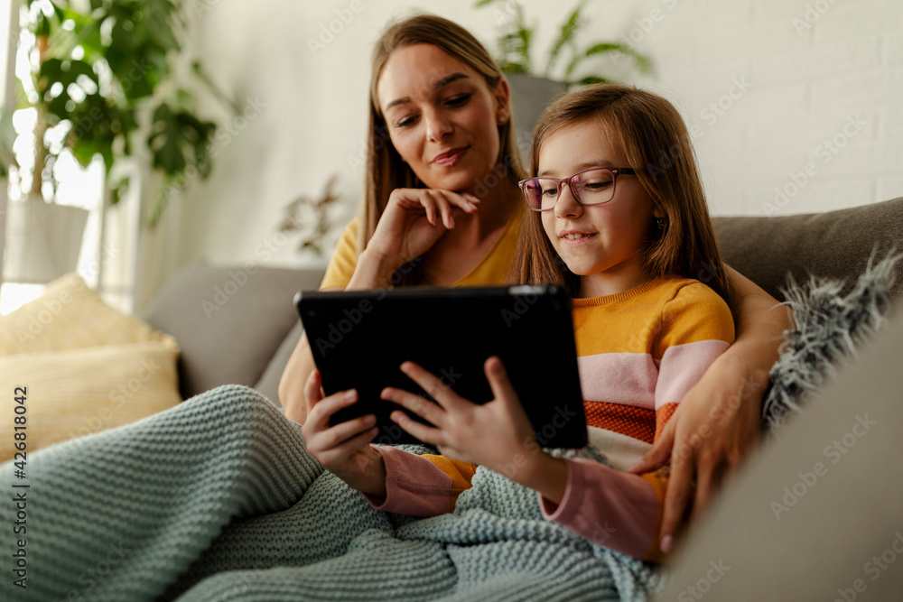 Smiling young Caucasian mother and small preschooler daughter sit relax on couch using tablet. Smiling young Caucasian mother and small preschooler daughter sit relax on couch using tablet.