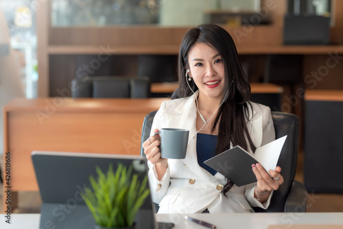 Asian businesswoman wearing headphone and holding coffee cup and book sitting relax the tablet is placed at the office table. Looking at camera.