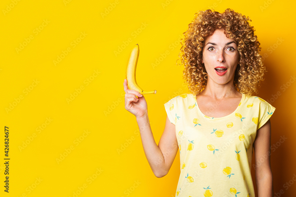 shocked surprised curly young woman holding a banana on a yellow background shocked surprised curly young woman holding a banana on a yellow background