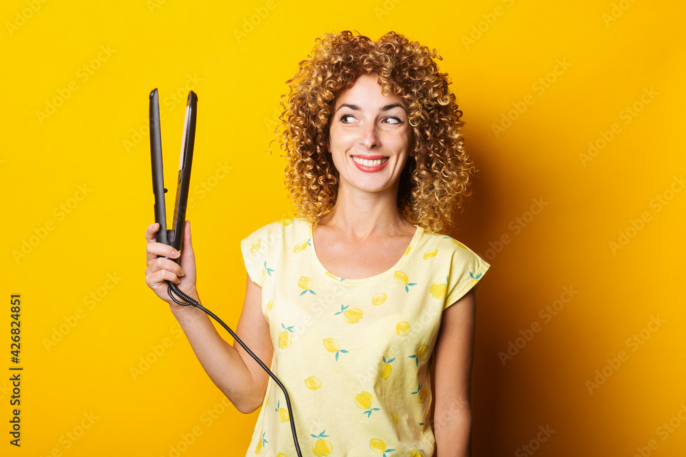 young woman with curly hair smiling looks at a hair straightener on a yellow background young woman with curly hair smiling looks at a hair straightener on a yellow background