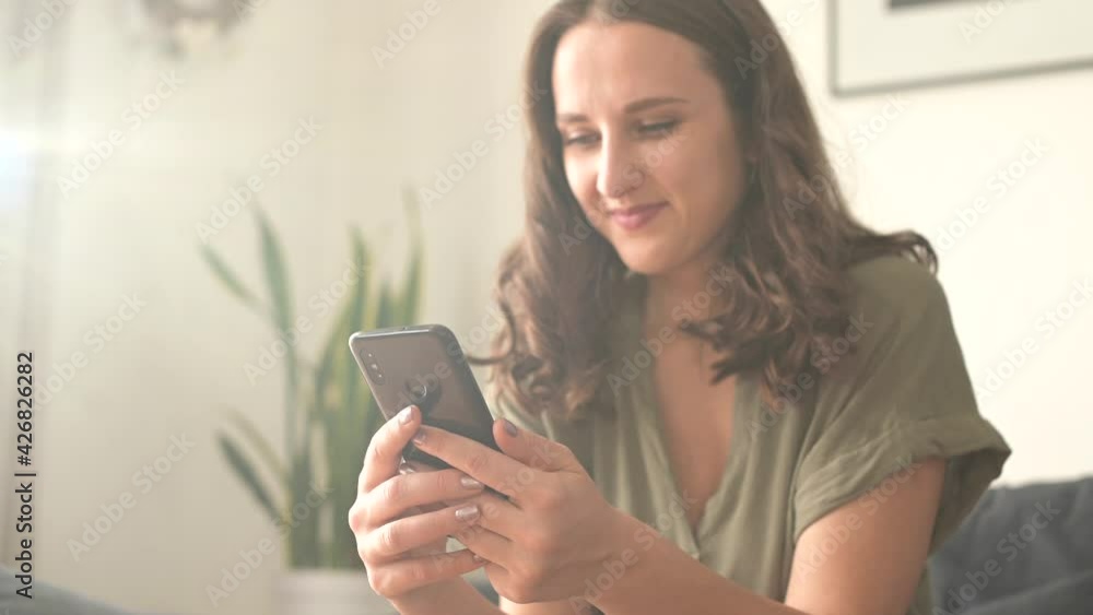Selective focuus at the phone in hands of female. Cheerful young woman enjoys chatting online, sits on the couch at home and messaging, scrolling news feed on the smartphone Selective focuus at the phone in hands of female. Cheerful young woman enjoys chatting online, sits on the couch at home and messaging, scrolling news feed on the smartphone