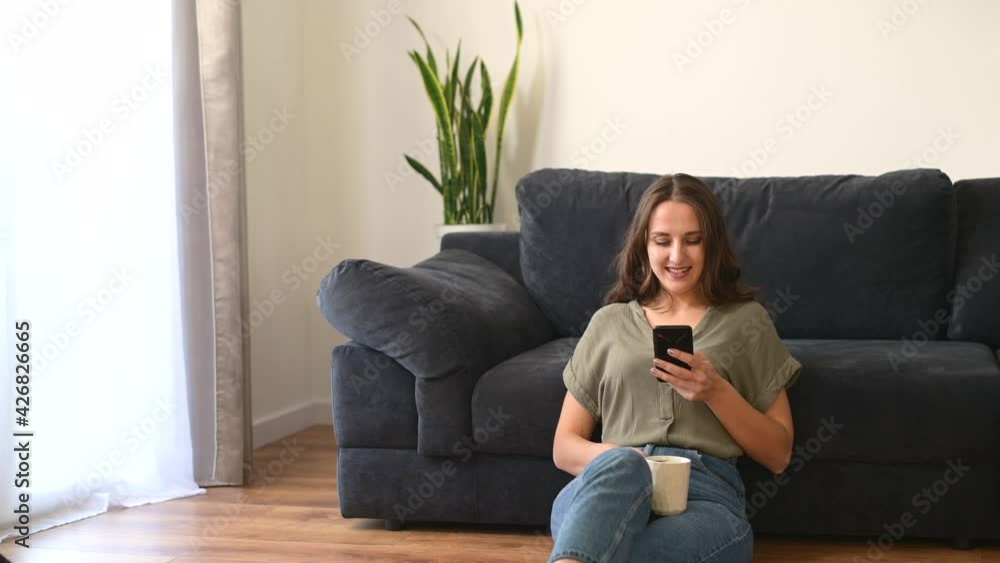 Pretty young woman using smartphone and drinking coffee sitting on the floor in cozy living room, smiling girl is messaging online on the phone, holds mug with hot drink, checks emails Pretty young woman using smartphone and drinking coffee sitting on the floor in cozy living room, smiling girl is messaging online on the phone, holds mug with hot drink, checks emails
