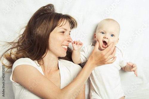 Young mother plays with her baby on a bed with a white sheet. Flat lay, top view