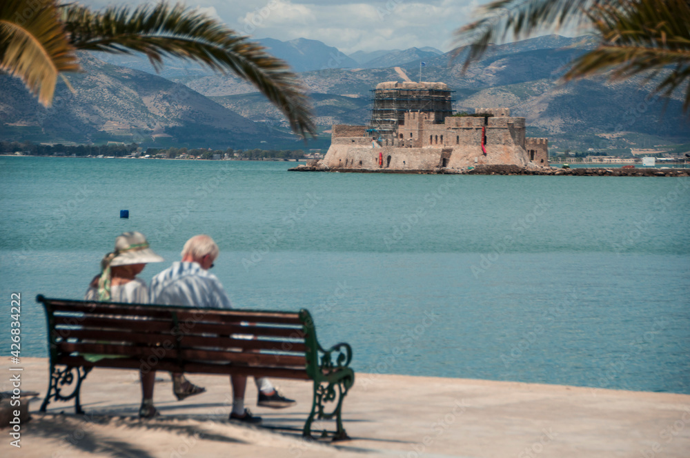 A senior couple sitting on a bench enjoying a view of Venetian Bourtzi Castle in Nafplio, Greece