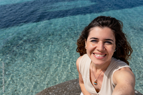 Self portrait or selfie photo of an attractive happy, smiling young woman on the beach with beautiful translucent sea on background