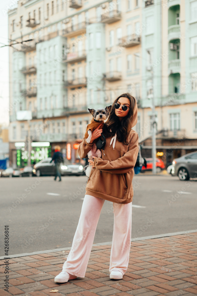 Fototapeta premium Vertical shot of a fashionable woman in sunglasses and stylish outfit hugging her small dog - york terrier, busy city street on a background.
