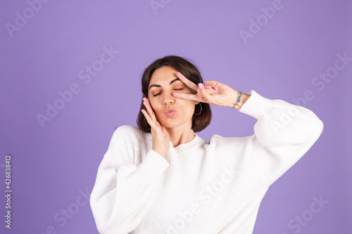Young brunette in white casual sweater isolated on purple background showing victory sign send air kiss