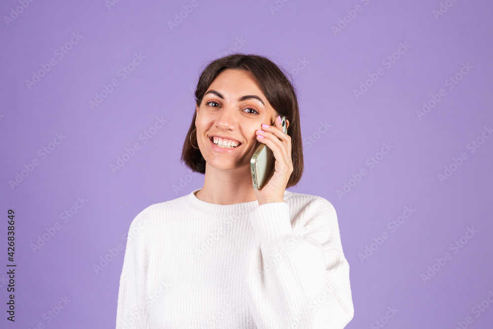 Young brunette in white casual sweater isolated on purple background having conversation with happy mood, talking positive on mobile phone, smiling Young brunette in white casual sweater isolated on purple background having conversation with happy mood, talking positive on mobile phone, smiling