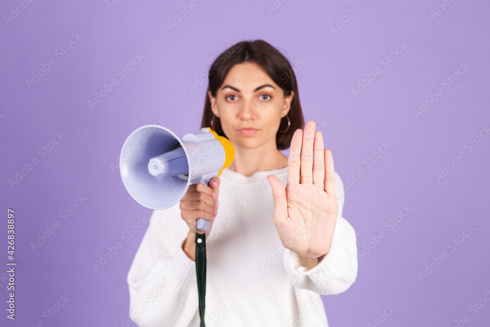 Young brunette in white casual sweater isolated on purple background unhappy serious with megaphone doing stop sing with palm of the hand. Warning expression Young brunette in white casual sweater isolated on purple background unhappy serious with megaphone doing stop sing with palm of the hand. Warning expression