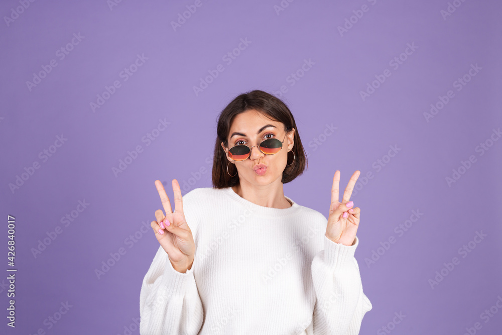 Young brunette in white casual sweater isolated on purple background smile to camera do peaceful gesture victory sign Young brunette in white casual sweater isolated on purple background smile to camera do peaceful gesture victory sign