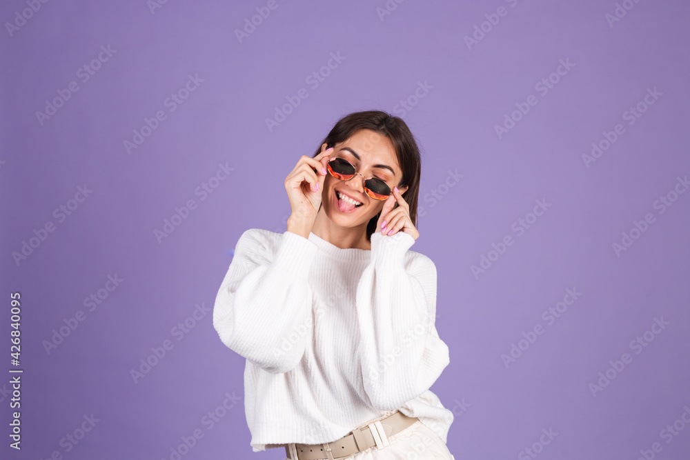 Young brunette in white casual sweater isolated on purple background in sunglasses happy funny playful shows tongue Young brunette in white casual sweater isolated on purple background in sunglasses happy funny playful shows tongue