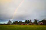 A cloudy landscape view of open field filled with grass with red cottages and rainbow in the sky in a rainy day during a outdoor visit