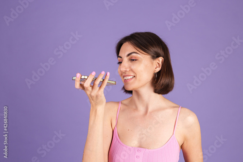 Young brunette in top isolated on purple background holding mobile phone recording audio voice message happy smile, having conversation
