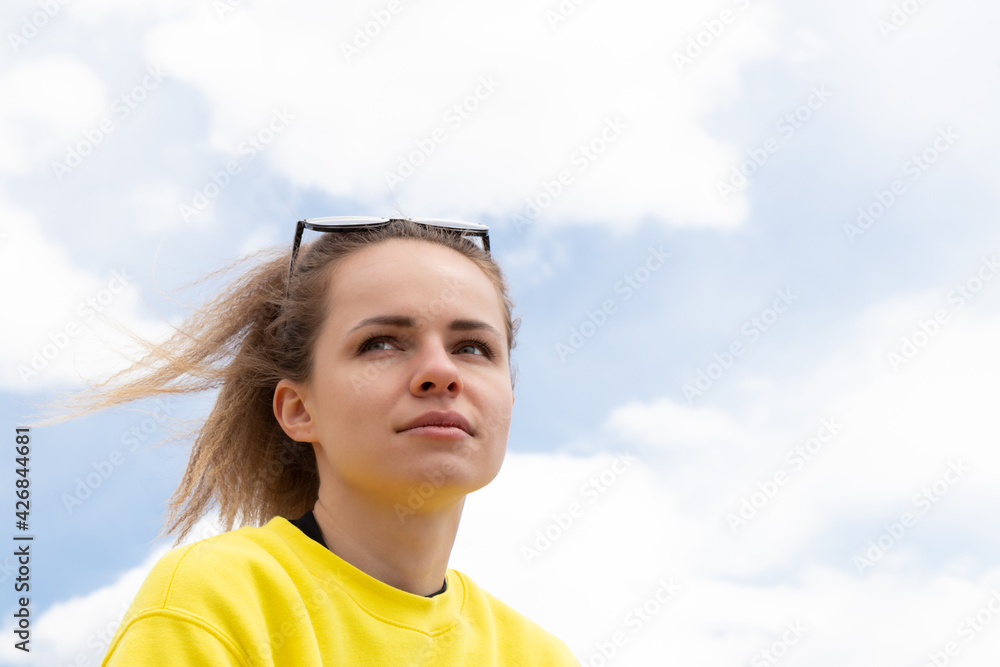 Reflective and authentic young girl wearing yellow sweatshirt looking in the distance sitting outside under blue sky and white clouds. Dreaming about positive future and enjoying life concept. Reflective and authentic young girl wearing yellow sweatshirt looking in the distance sitting outside under blue sky and white clouds. Dreaming about positive future and enjoying life concept.