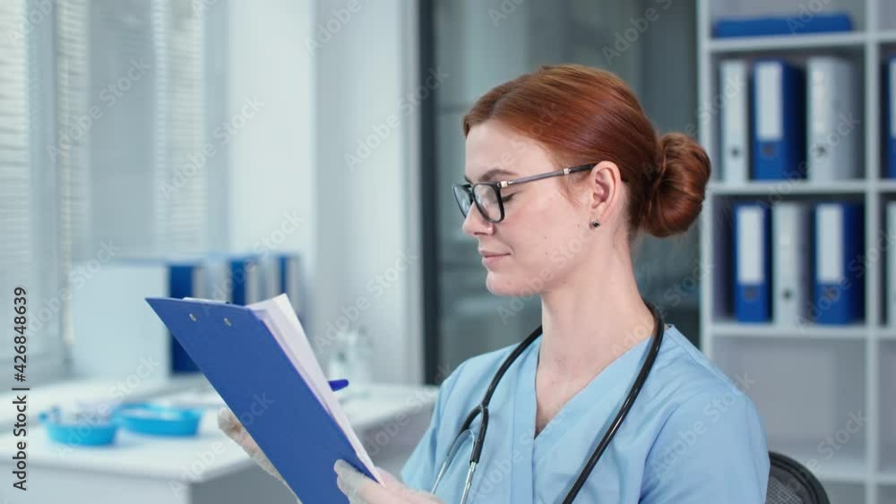 female physician in safety gloves writes notes in the clipboard, happy therapist in glasses and blue uniform looking at camera in hospital female physician in safety gloves writes notes in the clipboard, happy therapist in glasses and blue uniform looking at camera in hospital