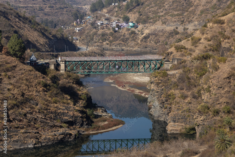 View of an Iron foot bridge over a river flowing with a village in the distant background and rocky cliffs on the sides in Kumaun region of UttaraKhand India on 12 January 2021